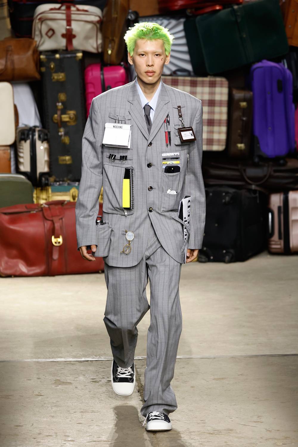 Modelo desfilando peças do verão 2025 da Moschino, usando terno risca de giz cinza, com aplicações de diversos materiais de escritório, como canetas, calculadoras, celular, óculos, etc.