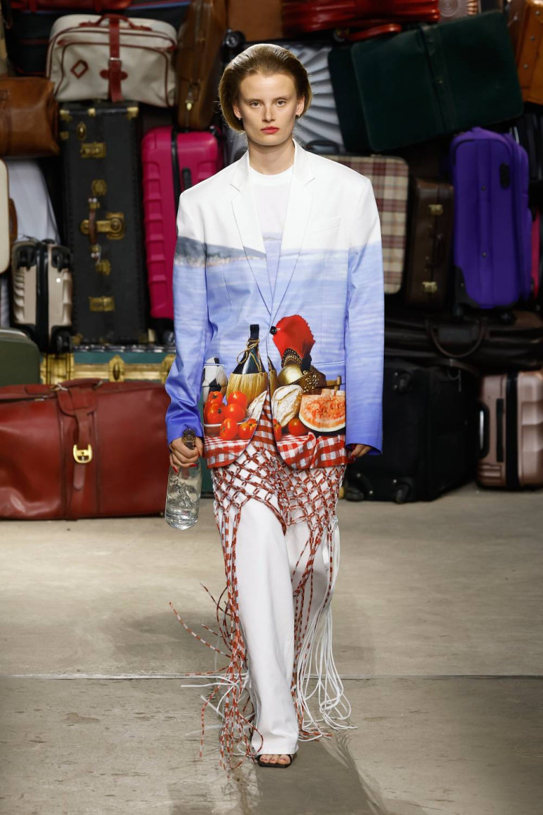 Modelo desfilando peças do verão 2025 da Moschino, usando terno com uma estampa em forma de mesa italiana com fundo do horizonte do mar, calça branca e uma saia de macramê vermelha sobre a calça. 