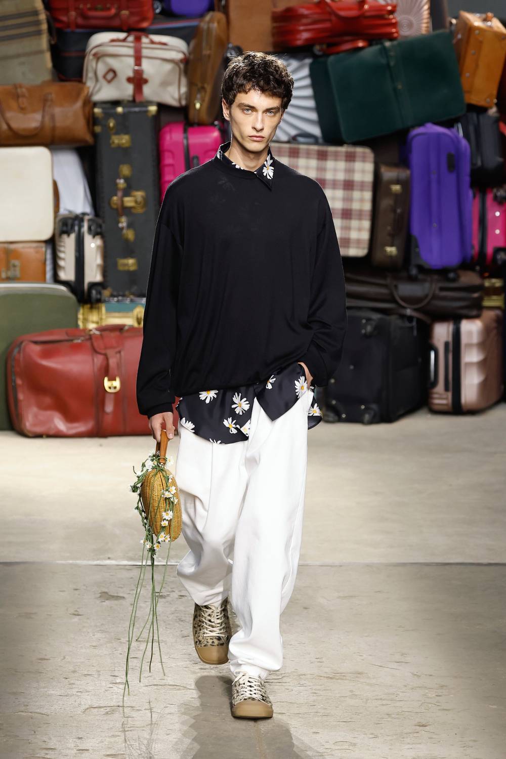 Modelo desfilando peças do verão 2025 da Moschino, usando camisa estampada de flores brancas sob suéter preto, calça ampla branca e tênis com estampa de onça.
