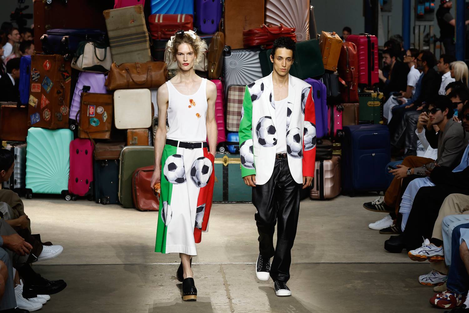 Dois modelos desfilando peças do verão 2025 da Moschino, a mulher usa uma regata branca manchada de molho de tomate e bola estampada sobre a bandeira italiana. O rapaz usa blazer com a mesma estampa e calça de couro preta.