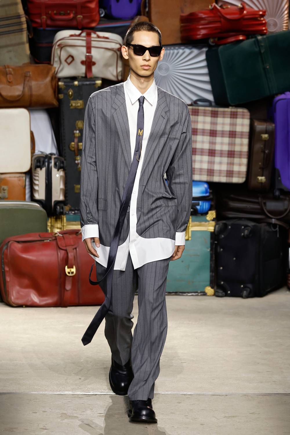 Modelo desfilando peças do verão 2025 da Moschino, com terno cinza, camisa branca, gravata superlonga e óculos de sol.