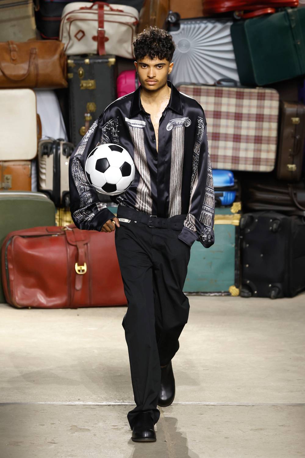 Modelo desfilando peças do verão 2025 da Moschino, usando camisa estampada com colunas gregas, bolsa de futebol usada como bolsa e calça preta social.