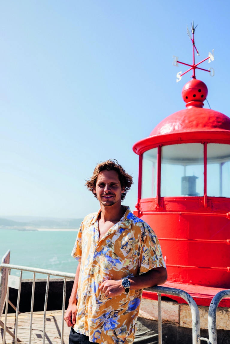 Nic von Rupp posa a frente de um farol, com o mar ao fundo. O surfista está usando uma camisa estampada colorida.