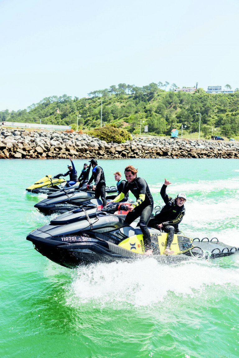 Três jet skis em alto-mar, com Nic von Rupp pilotando o que está em primeiro plano. Em cada jet ski, há dois tripulantes a bordo. Ao fundo, é possível ver um pouco das pedras e da paisagem natural, cheia de árvores, de Nazaré, em Portugal.
