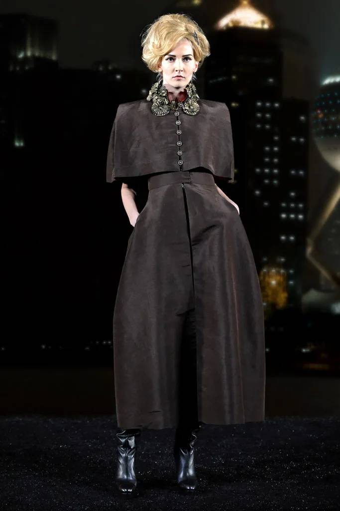 Uma modelo loira usa peça da coleção Métiers d'Art 2010: macacão preto, com ombros retos e pernas amplas, em formato arredondado. No pescoço, um colar com amplas flores de metal prateado e, nos pés, botas de couro preto.