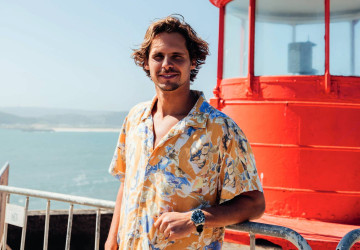 Nic von Rupp posa a frente de um farol, com o mar ao fundo. O surfista está usando uma camisa estampada colorida.